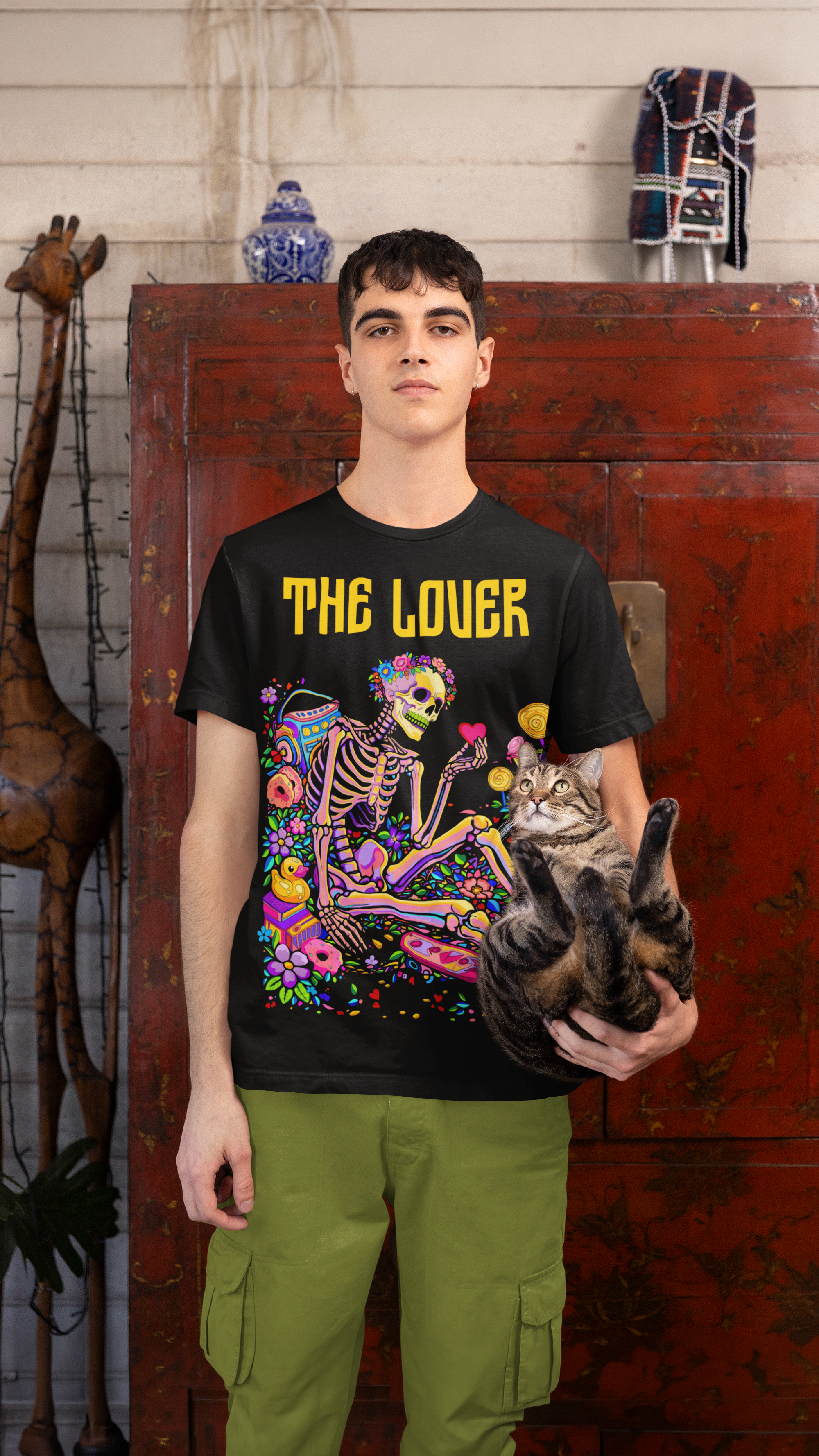 Person wearing a black t-shirt with colorful skeleton design and 'THE LOVER' text, holding a cat in front of a wooden cabinet.